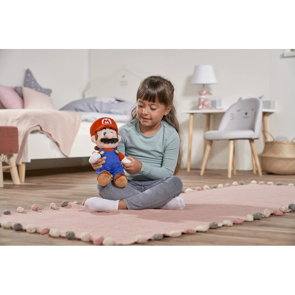Een jong meisje met lang bruin haar zit gekruist op een roze kleed, met een Mario knuffel in een slaapkamer.