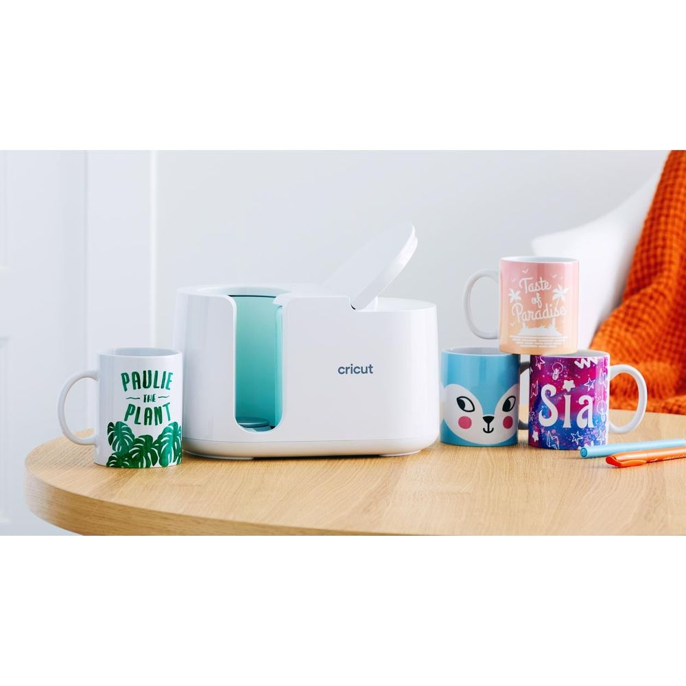 Una prensa para tazas Cricut blanca con cuatro tazas decoradas al lado sobre una mesa de madera.