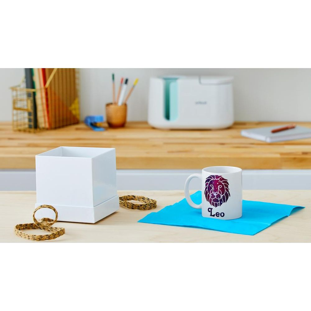 Caja blanca, taza Leo con diseño de león, sobre alfombrilla azul. Dispositivo al fondo.