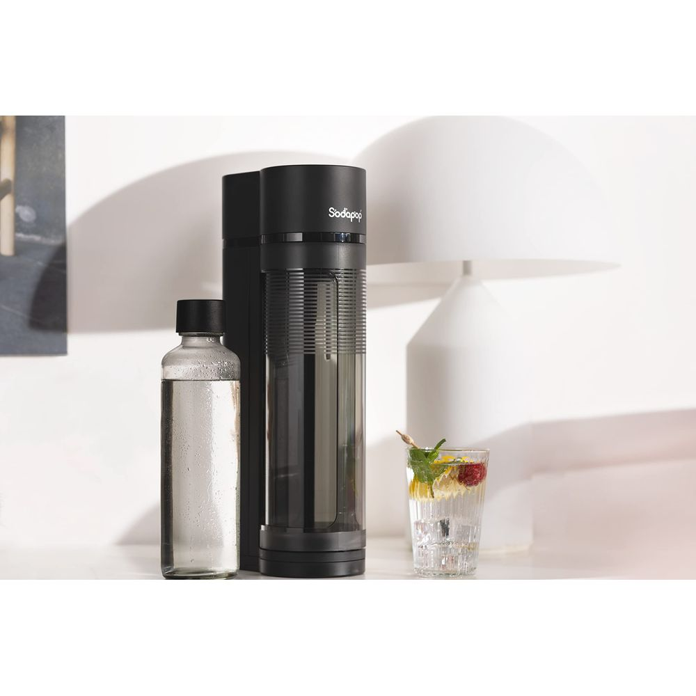 Ein schwarzer Sodastreamer steht neben einer Wasserflasche und einem Glas mit Getränk.