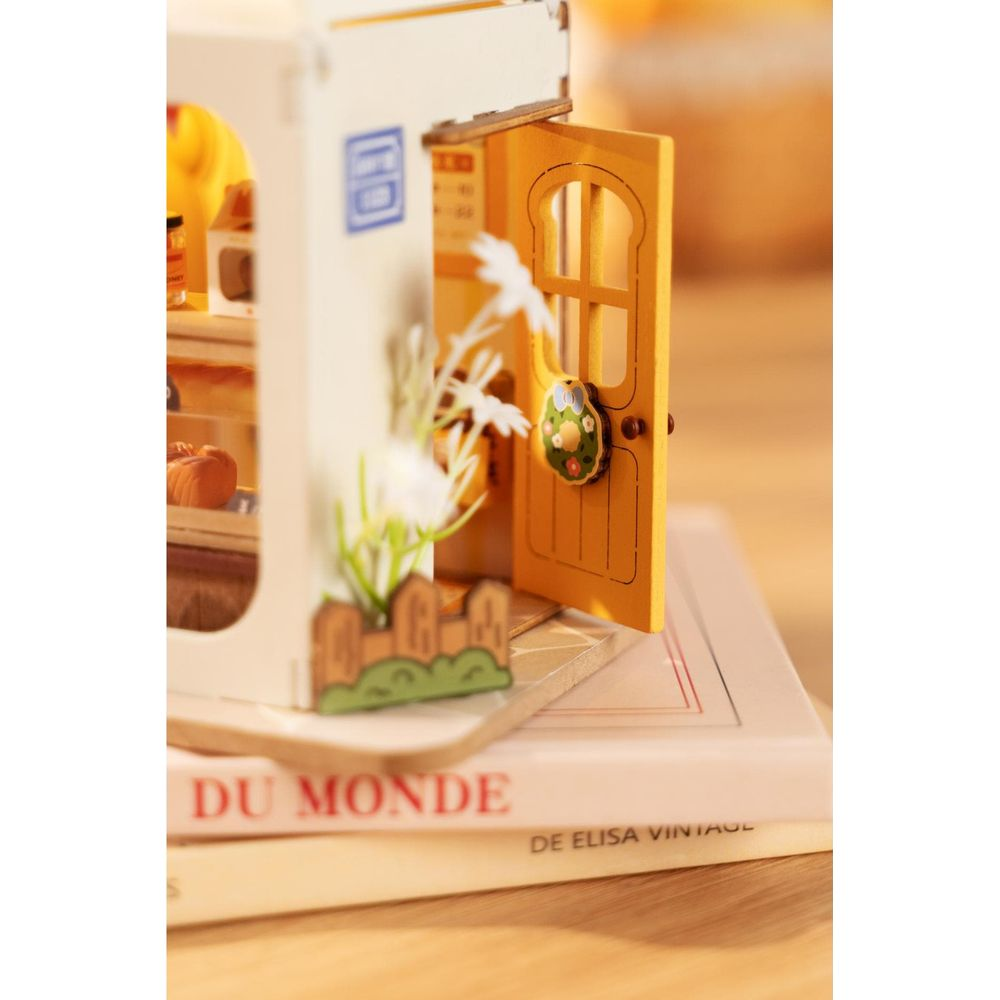 Casa in miniatura con porta gialla, fiori e espositore di pane sui libri.