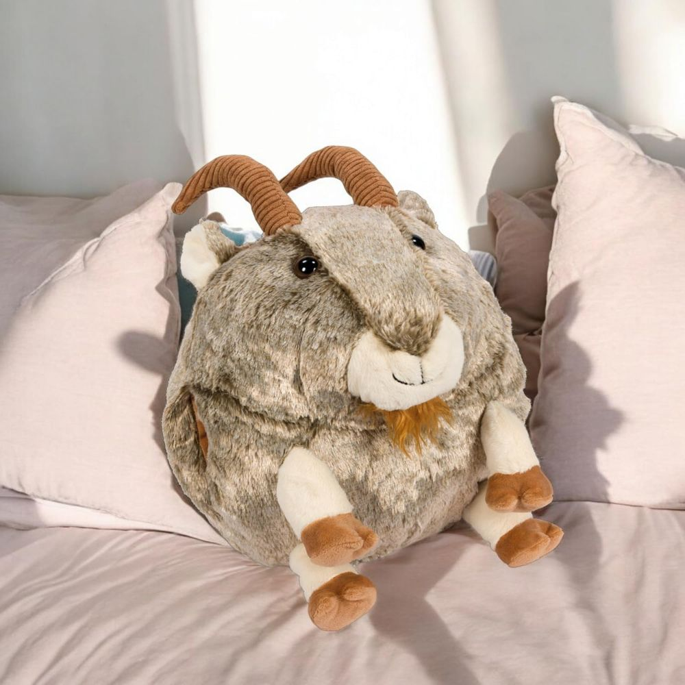 Ein flauschiges Ziegen-Plüschtier sitzt auf einem Bett zwischen rosa Kissen.