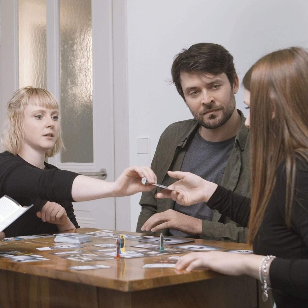 Drei Personen spielen ein Brettspiel an einem Holztisch mit vielen Karten und Spielfiguren.