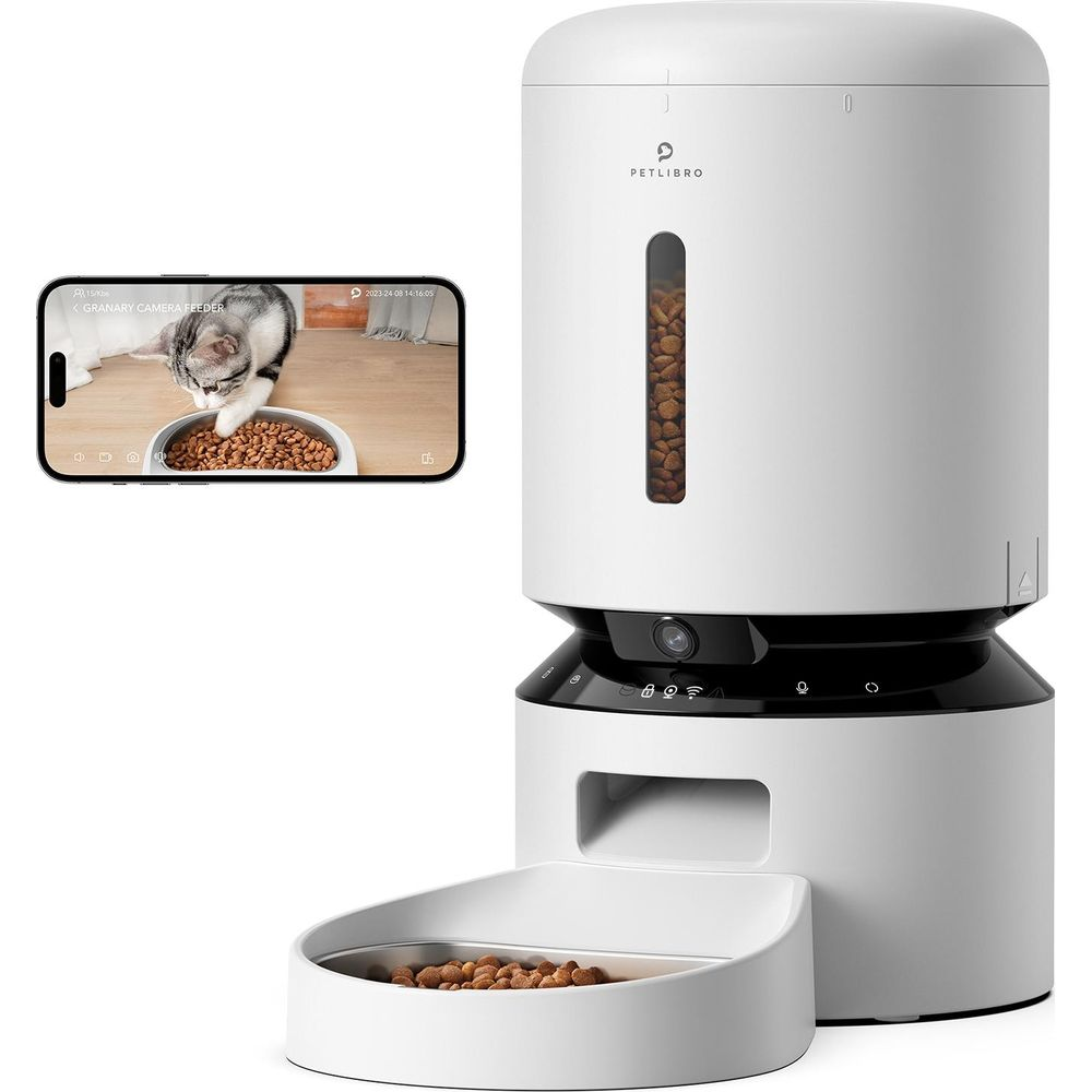 Un comedero blanco automático junto a la pantalla de un teléfono que muestra un gato comiendo.