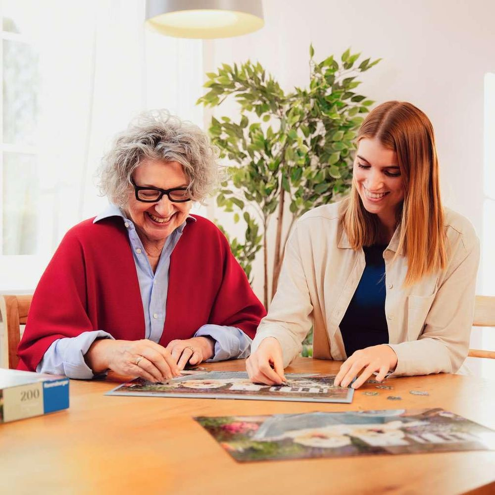 Due donne sorridenti completano un puzzle su un tavolo di legno con una pianta.