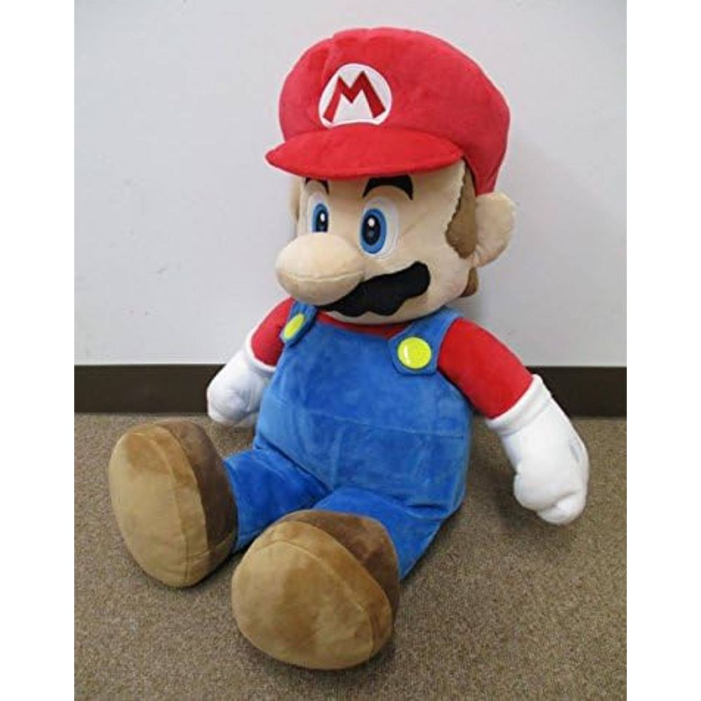 Un peluche di Mario seduto su un tappeto. Indossa un cappello rosso e una salopette blu.