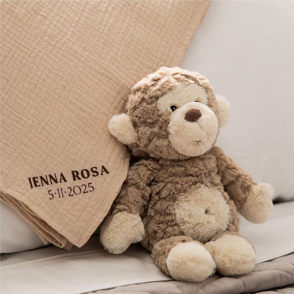 Ein brauner Teddybär sitzt neben einer beige Decke mit Namen und Datum.