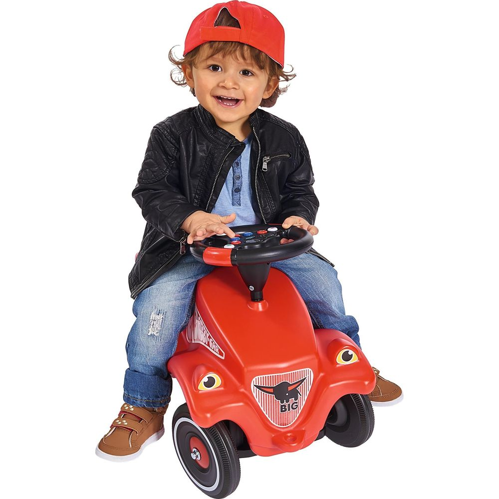 Ein Kleinkind in Lederjacke und roter Kappe sitzt auf einem roten Spielzeugauto.