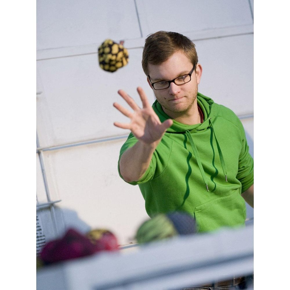 Mann im grünen Hoodie wirft gelbes Objekt mit schwarzen Flecken. Hand ausgestreckt.