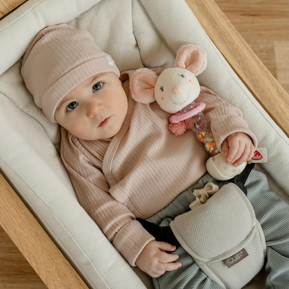 Un bimbo con cappello e tutina beige giace in una cesta, tenendo un sonaglio a forma di topo.