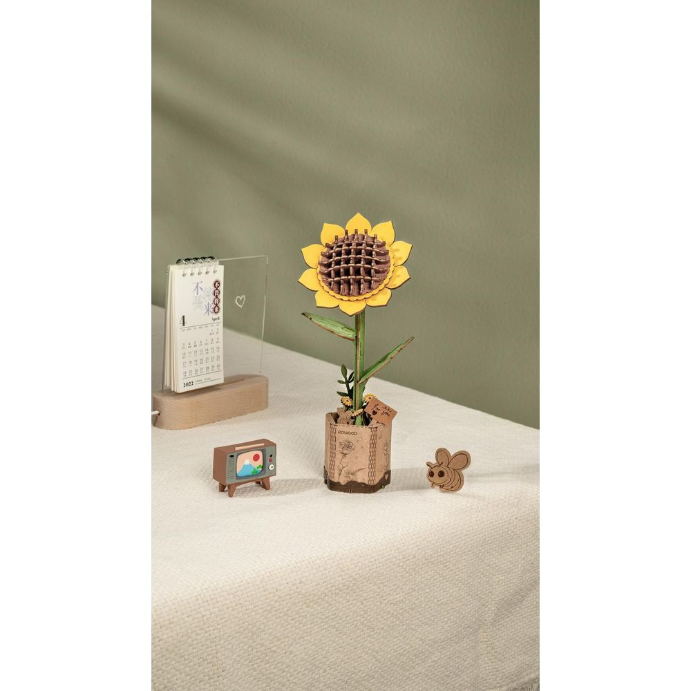 Drewniany model słonecznika stoi w doniczce, obok kalendarza i mini telewizora.