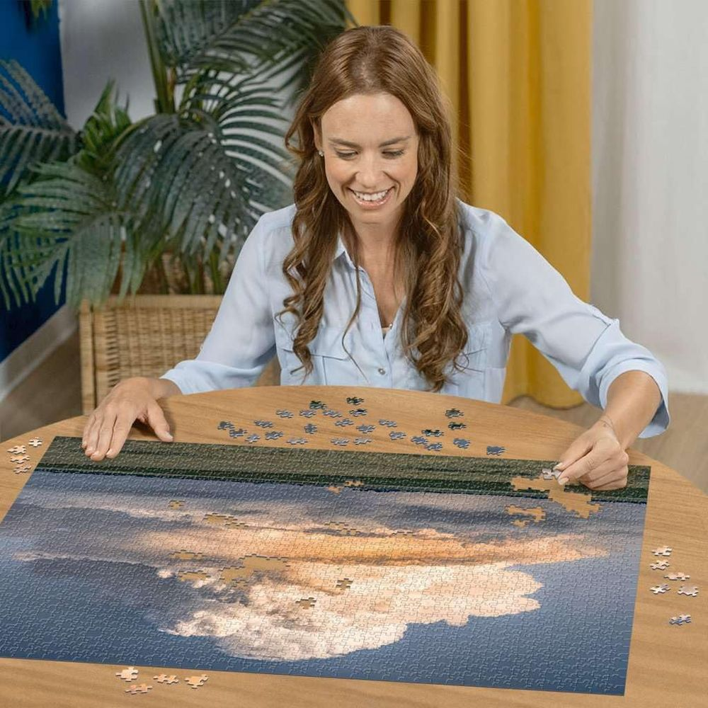 Donna che assembla un puzzle di un tramonto nuvoloso sull'acqua su un tavolo rotondo.