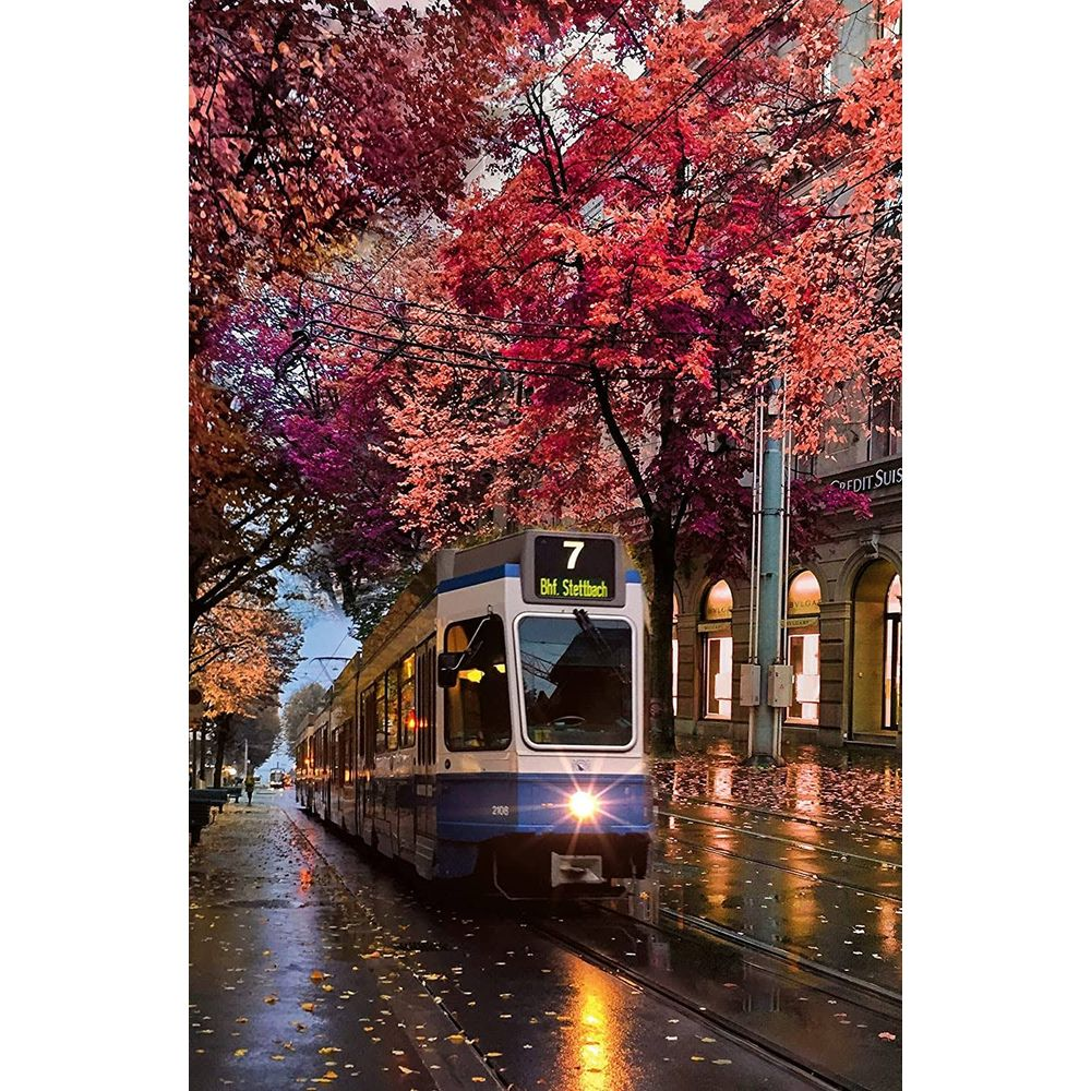 Niebiesko-biały tramwaj jedzie po mokrych torach, otoczony drzewami z czerwonymi liśćmi.