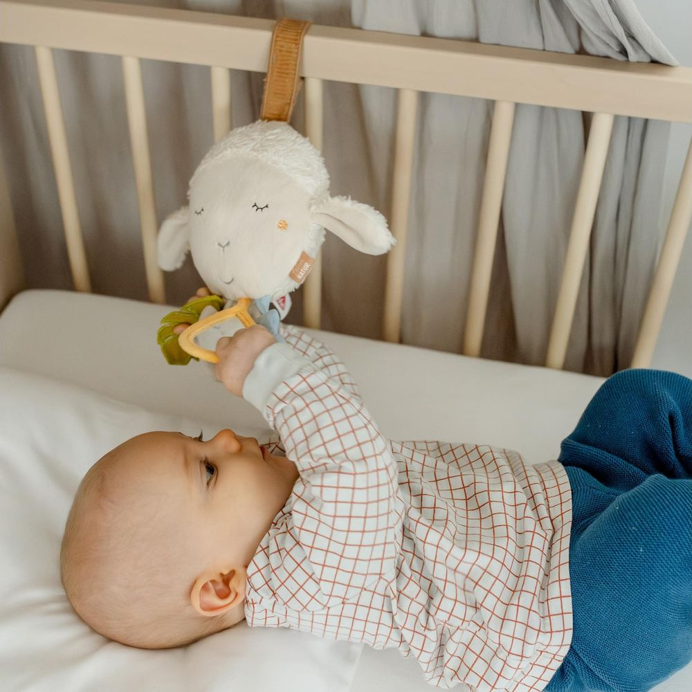 Ein Baby in einem karierten Oberteil spielt mit einem Schaf-Greifling in einem Kinderbett.