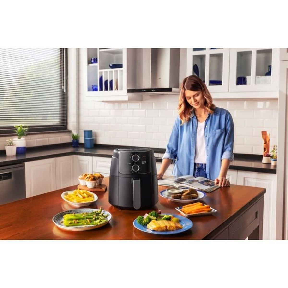 Kobieta w kuchni czyta książkę kucharską obok frytkownicy beztłuszczowej i gotowych potraw.