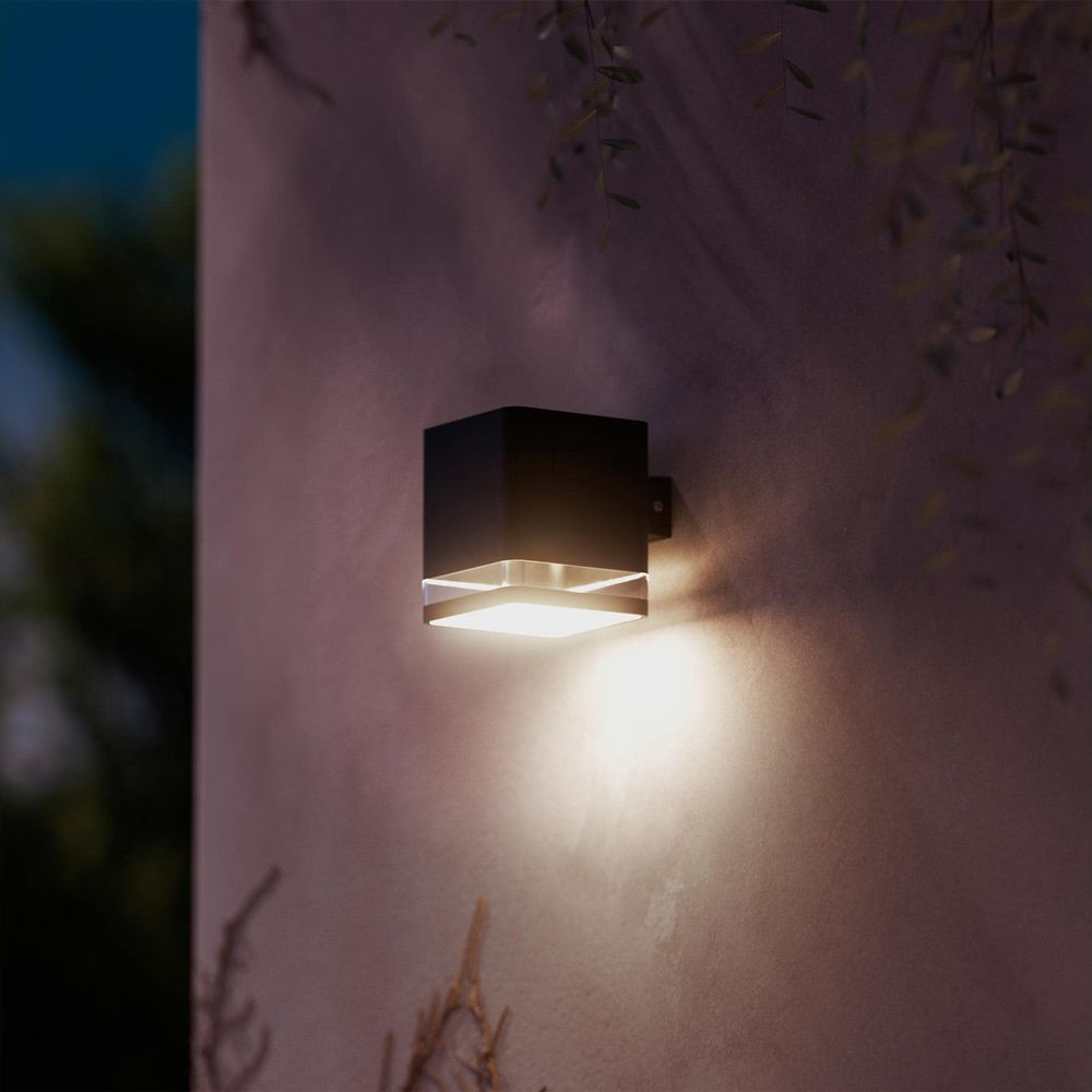 Ein schwarzer, würfelförmiger Außenwandleuchter leuchtet in der Dämmerung warm auf einer texturierten Wand.