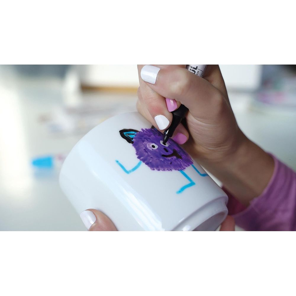 Ręka rysuje fioletowego potwora na białym kubku czarnym markerem.