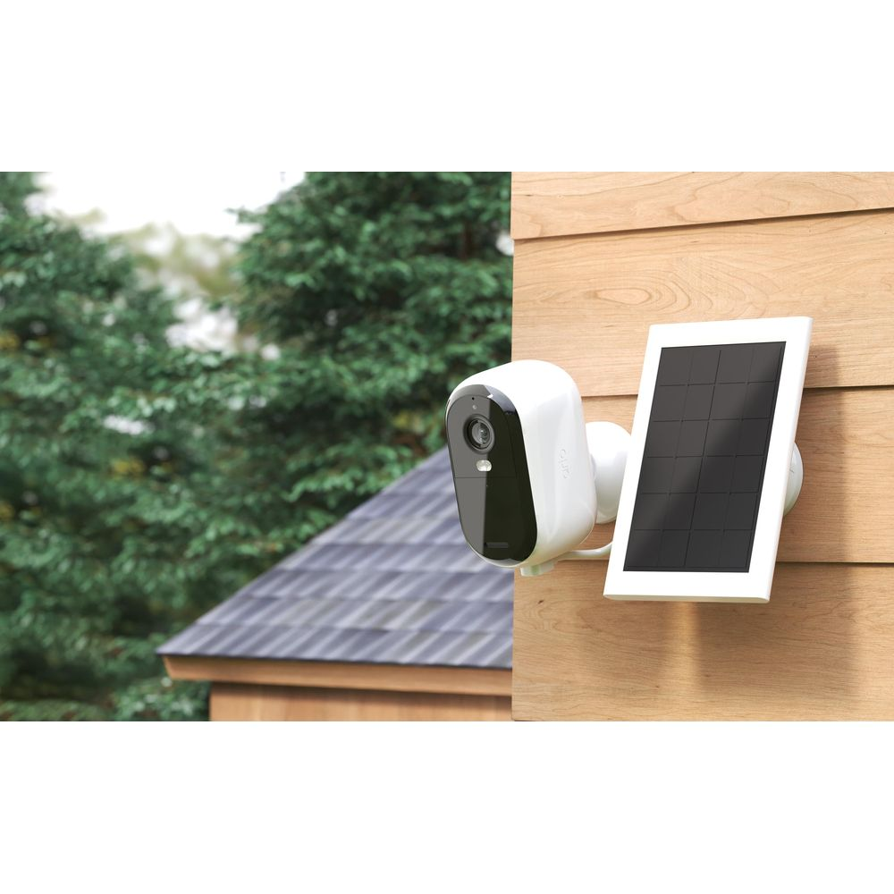 Eine weiße Überwachungskamera mit Solarpanel an einer Holzwand, Bäume im Hintergrund.