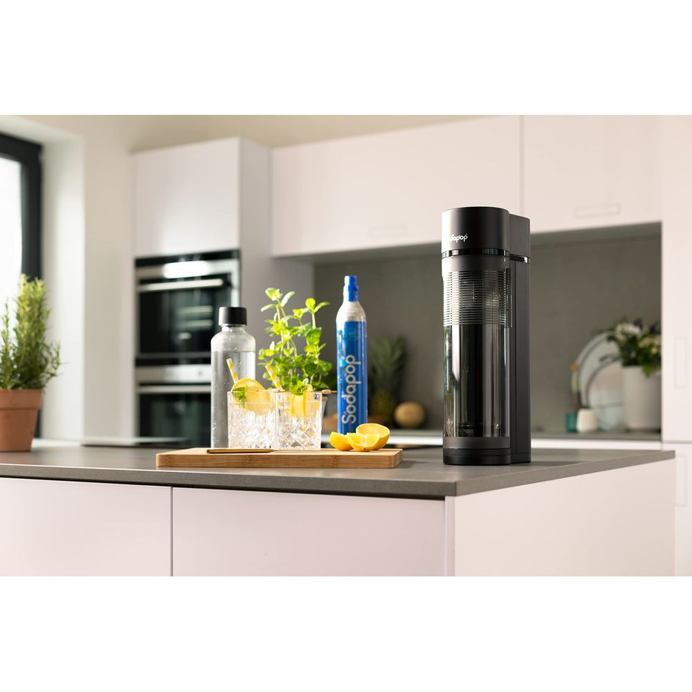Ein Soda-Maker steht auf einer Küchentheke neben Gläsern mit Getränken und Zitrone.