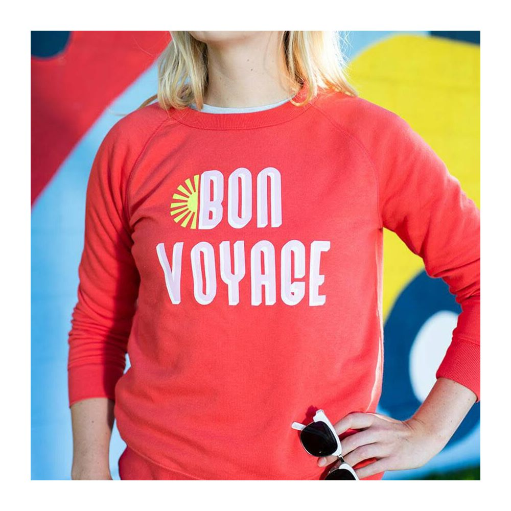 Eine Frau trägt ein rotes Sweatshirt mit „BON VOYAGE“ auf einer bunten Wand.