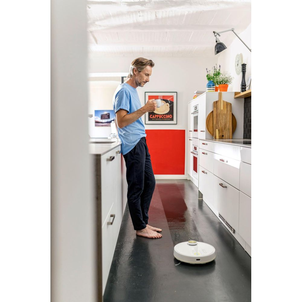 Un hombre con café en la mano está en una cocina moderna, un robot aspirador limpia el suelo.