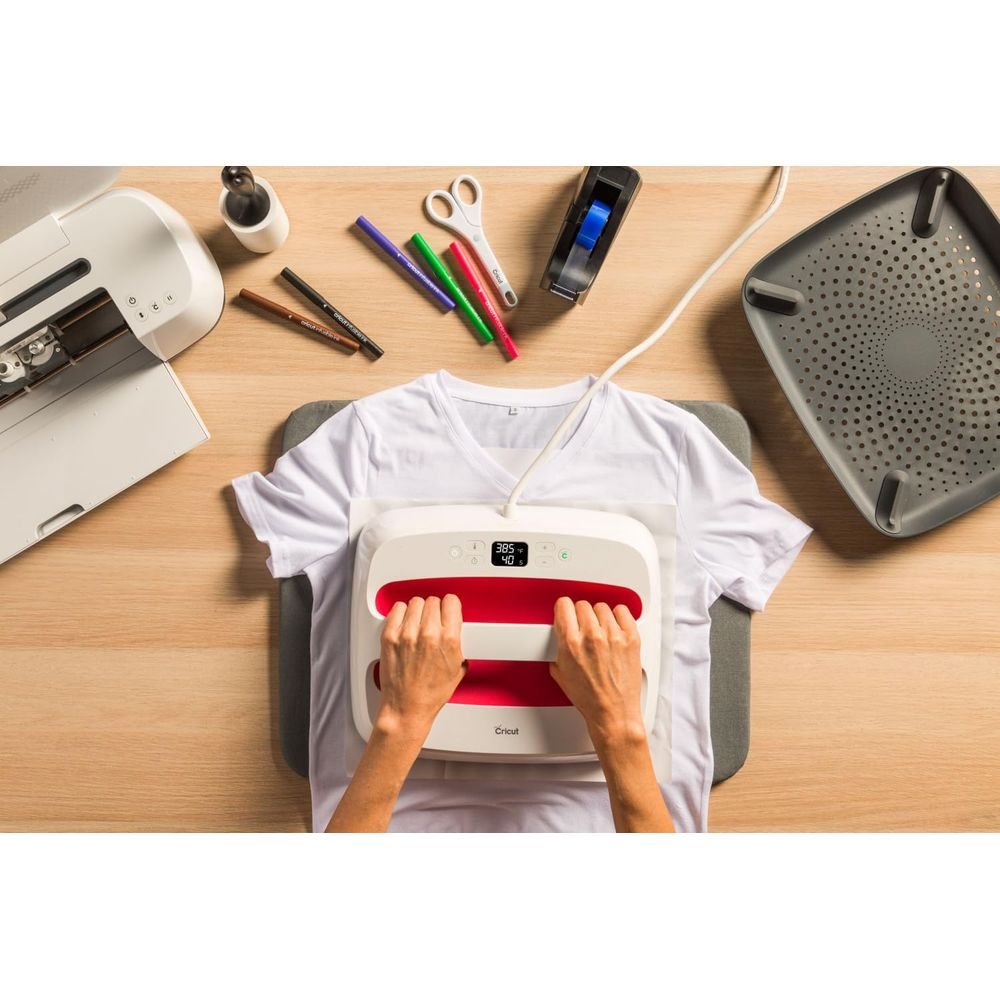 Hände bedienen eine Heißpresse auf einem weißen T-Shirt, Werkzeuge liegen daneben.