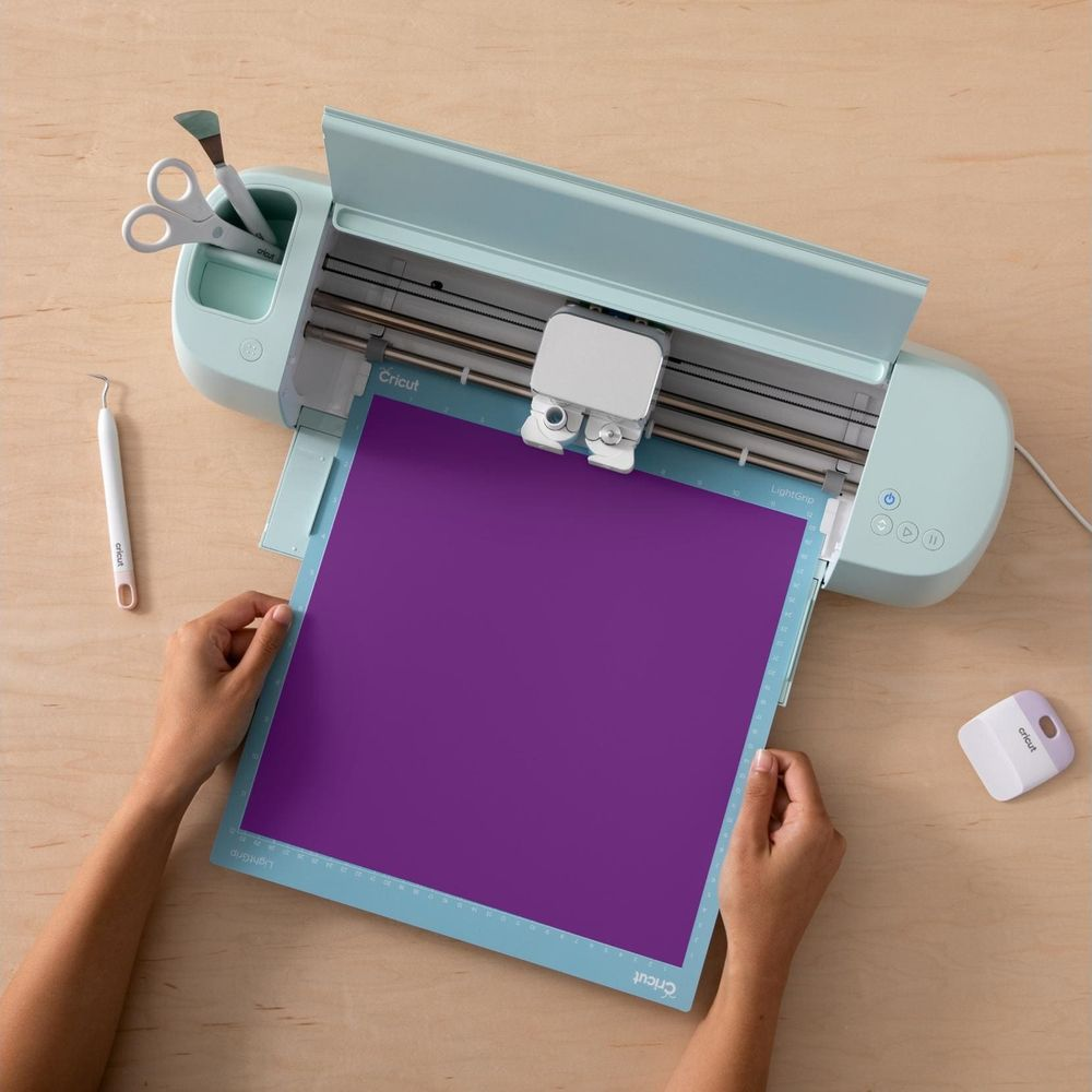 Hände legen lila Papier auf hellblaue Schneidematte neben Cricut-Maschine und Werkzeugen.
