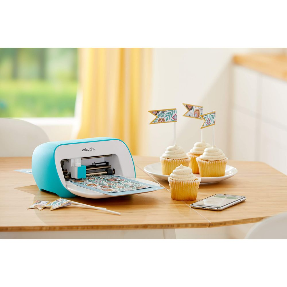 Une machine Cricut Joy turquoise avec une feuille de papier à motifs est sur une table en bois à côté de cupcakes.