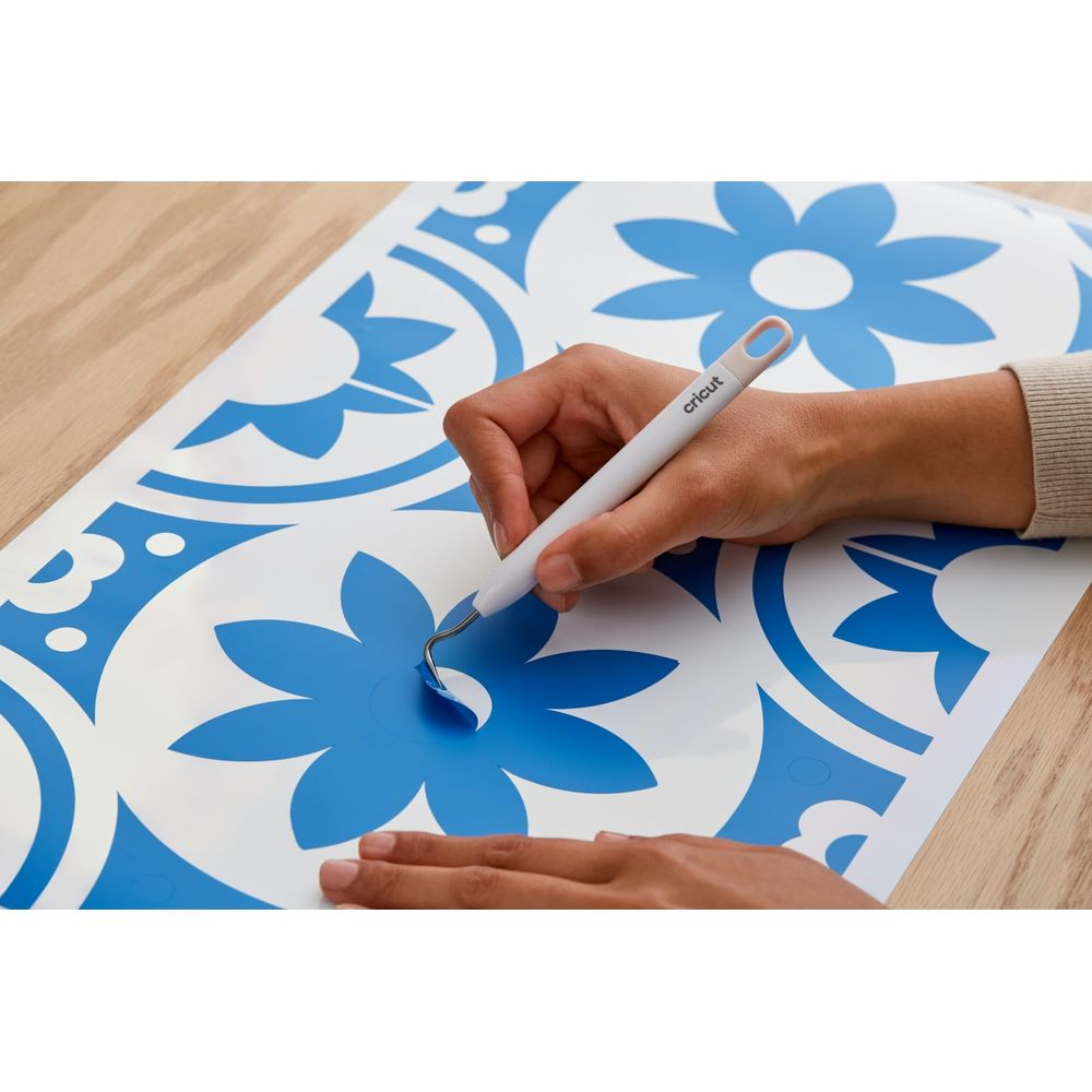 Eine Hand benutzt ein weißes Werkzeug, um blaue Vinylfolie von einer floralen Vorlage zu lösen.