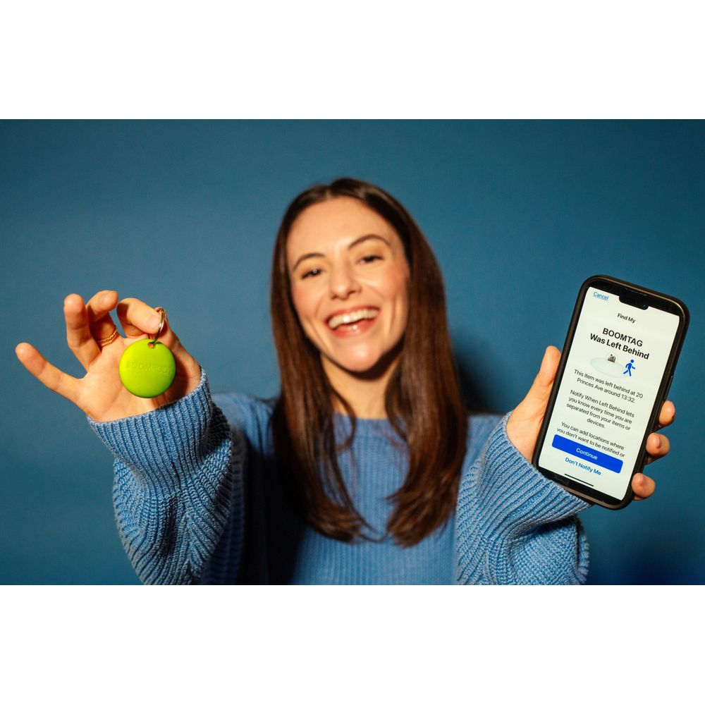 Uśmiechnięta kobieta trzyma zielony BOOMTAG i telefon z aplikacją 'Find My' z alertem o pozostawionym przedmiocie.