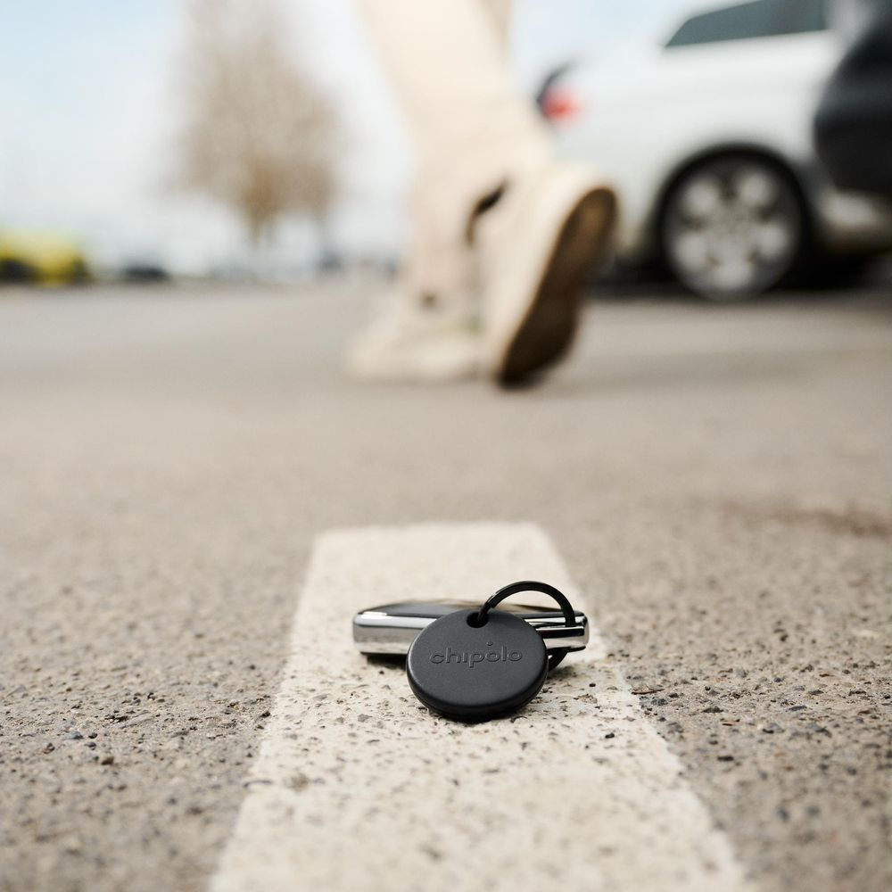 A black Chipolo keychain lies on the ground near cars.