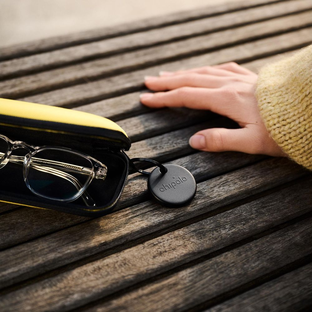 A hand rests on a wooden table next to glasses in a case and a Chipolo tracker.