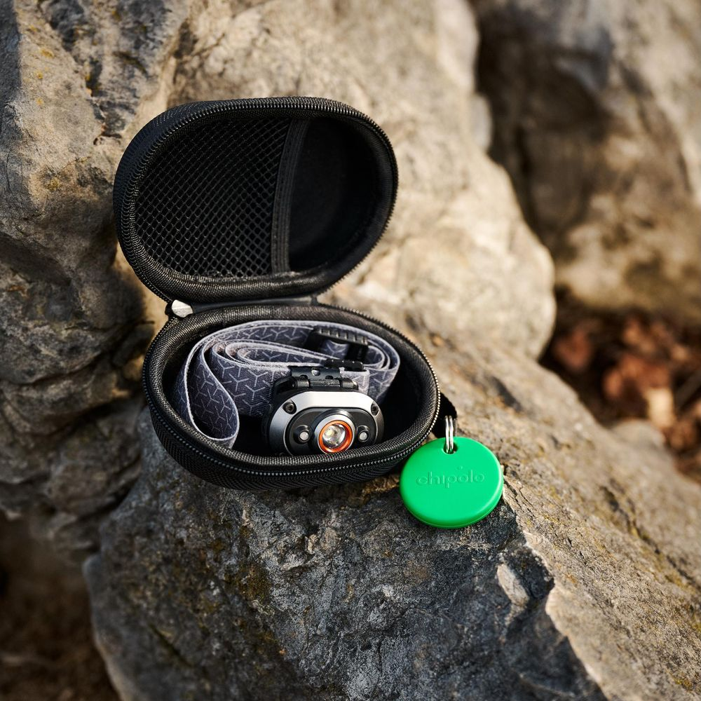 A headlamp inside a black case with a green Chipolo tracker on rocks.