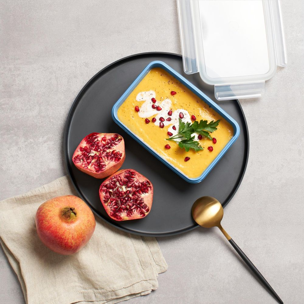 Aufnahme von oben: Orangensuppe in blauem Behälter mit Granatäpfeln, Löffel und Serviette auf grauem Untergrund.