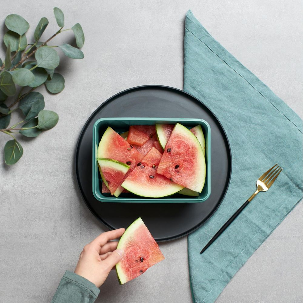 Eine Hand hält ein Stück Wassermelone neben einem Behälter voller Melonenstücke.