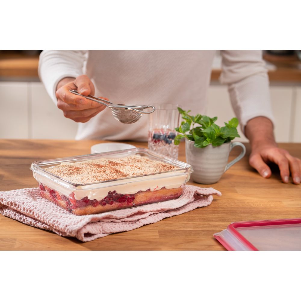 Eine Person bestäubt einen Kuchen mit Kakao aus einem Sieb. Ein Glas mit Beeren steht daneben.