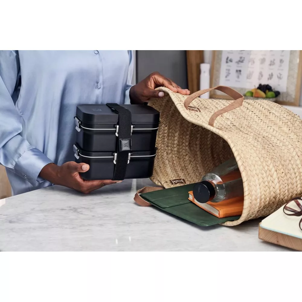 Eine Person hält eine schwarze Brotdose neben einer Strohtasche mit Flasche und Buch.