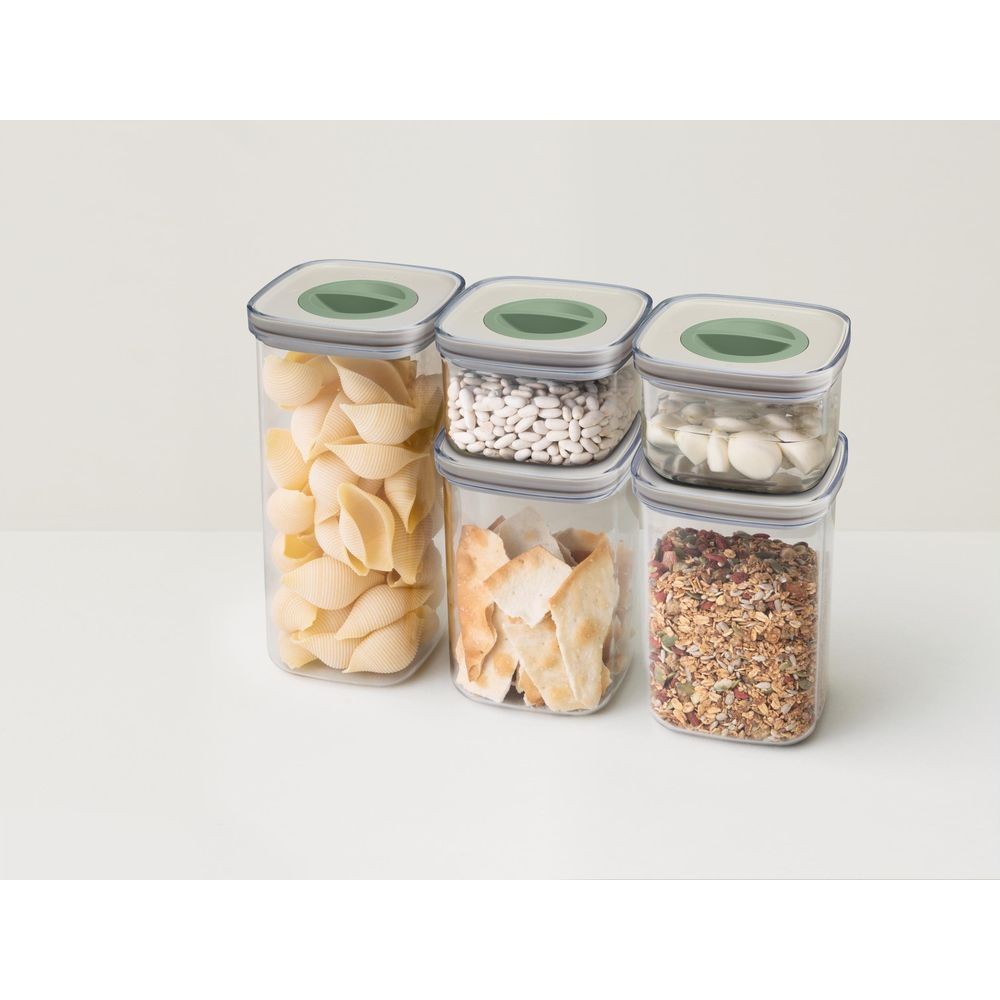 Five clear food storage containers with green lids, holding pasta, beans, bread, garlic, and granola.
