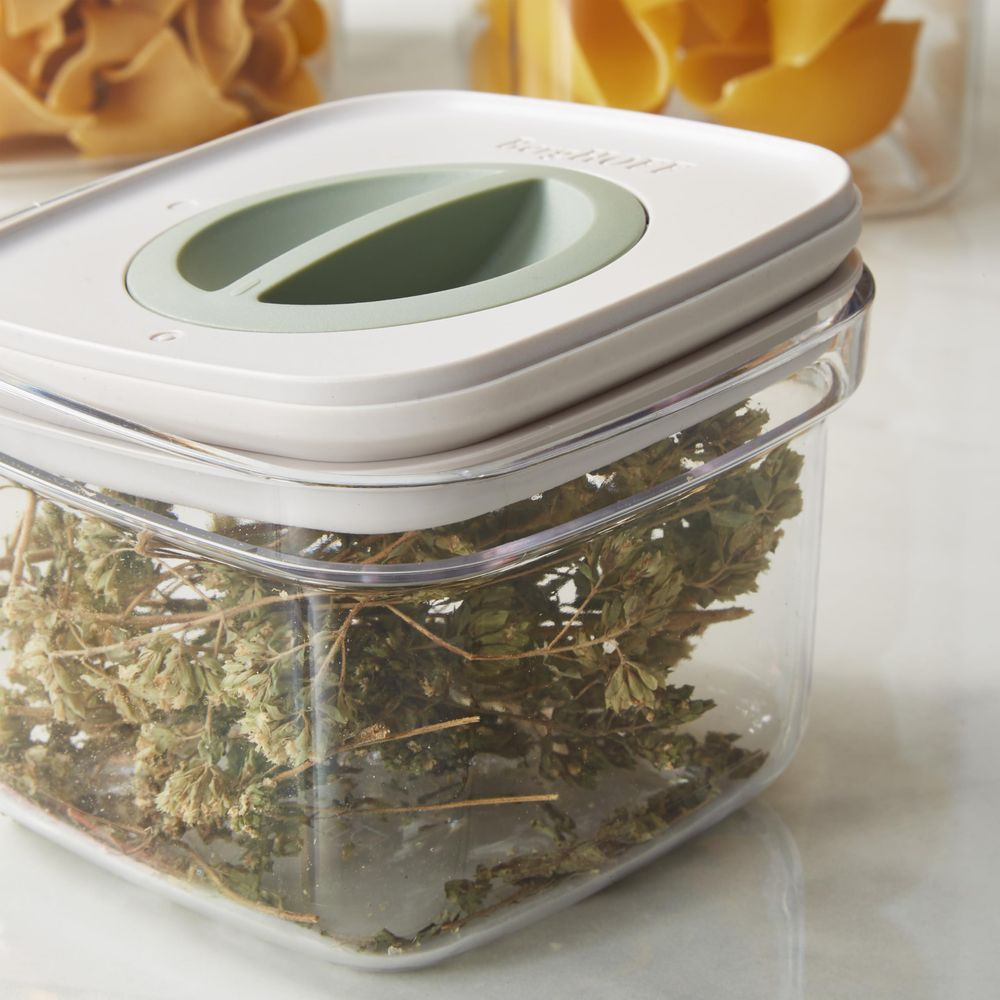 A clear container with a white lid and green handle, filled with dried herbs.