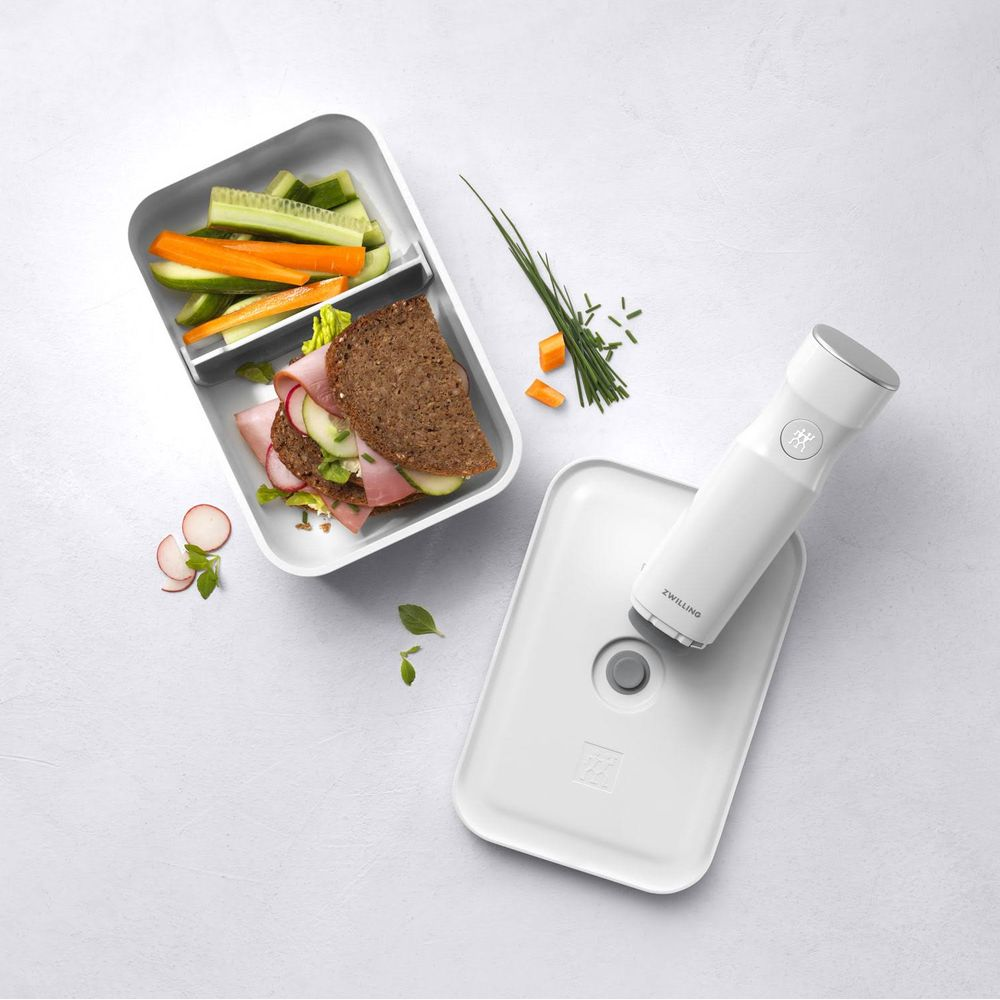 Un contenitore bianco per il pranzo con un panino, bastoncini di verdure e un sottovuoto.
