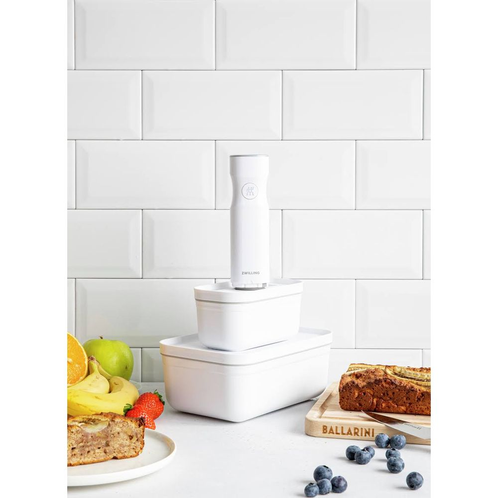 Contenitori bianchi per alimenti con sigillatore sottovuoto, pane, frutta e bacche su un tavolo.