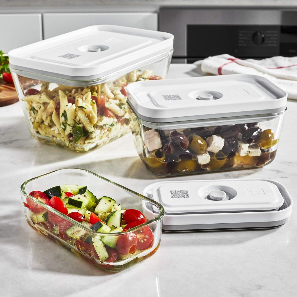 Tres recipientes de vidrio con comida en encimera de mármol: ensalada de pasta, aceitunas con feta y ensalada de pepino y tomate.