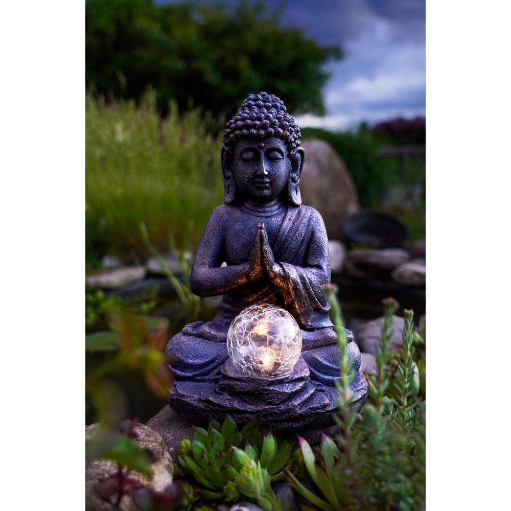 Buddha-Statue mit leuchtender Kugel im Garten, umgeben von Steinen und Pflanzen.