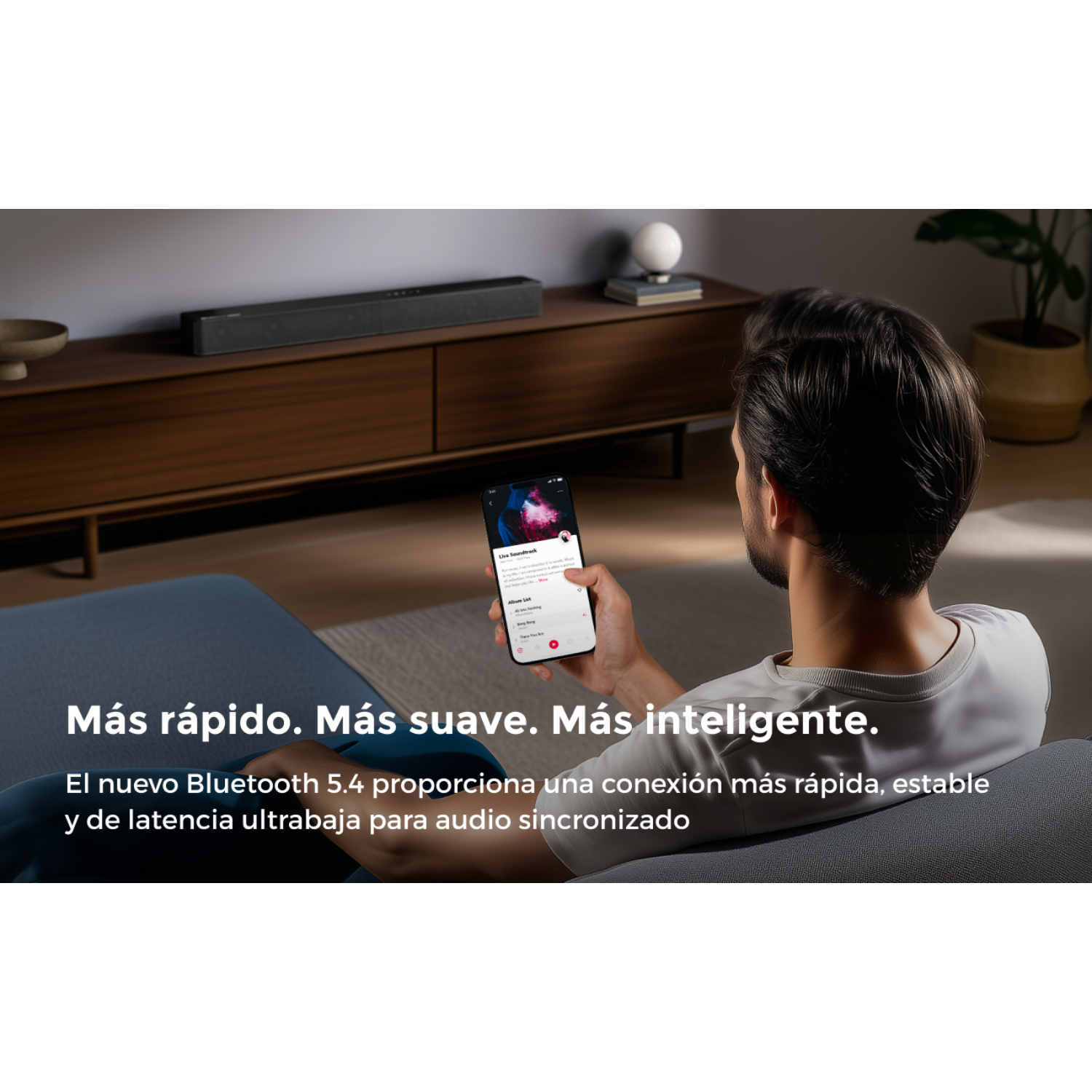 Hombre en sofá sostiene smartphone, barra de sonido detrás.