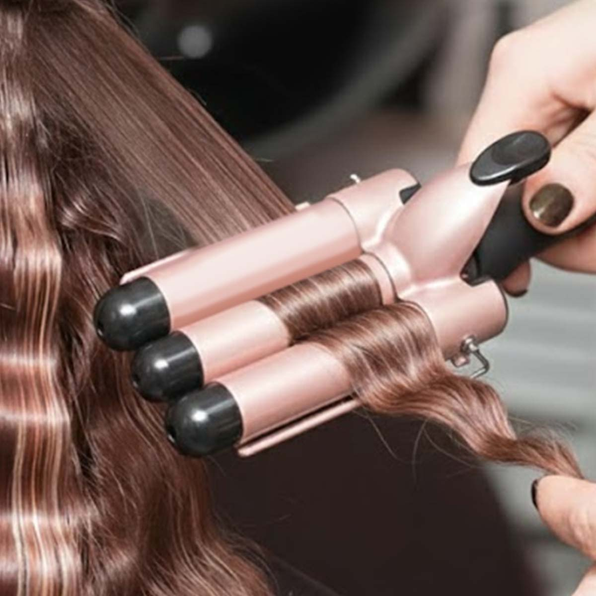 Eine Person stylt braunes Haar mit einem roségoldenen Dreifach-Lockenstab.