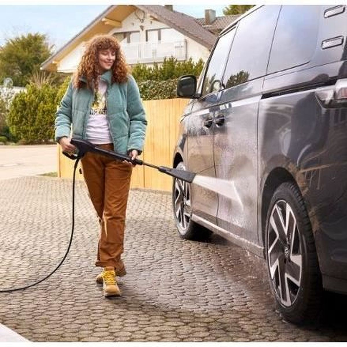 Una donna lava un'auto scura con un'idropulitrice su un vialetto pavimentato.
