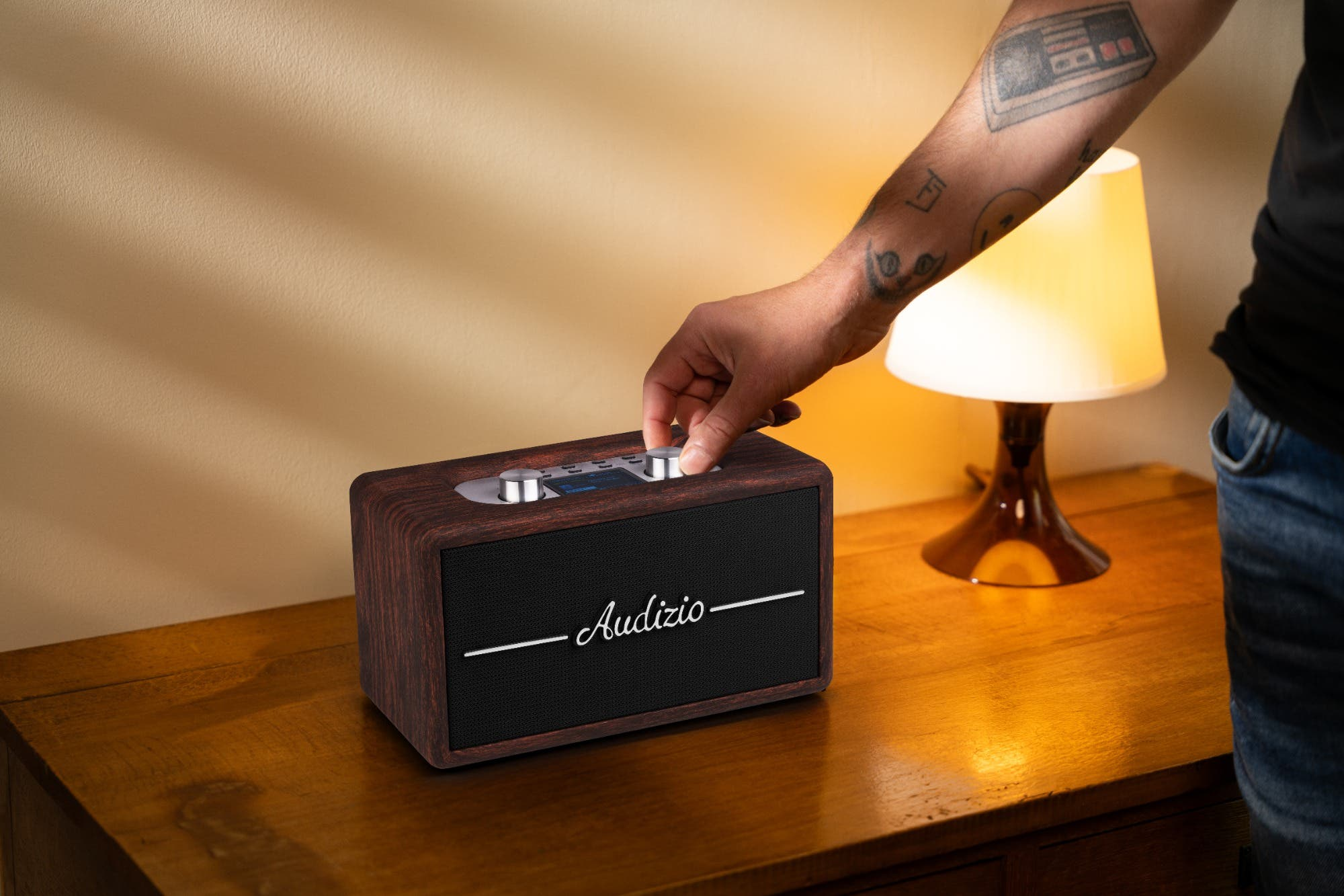 Una mano gira un botón de una radio de madera en una mesita de noche junto a una lámpara.