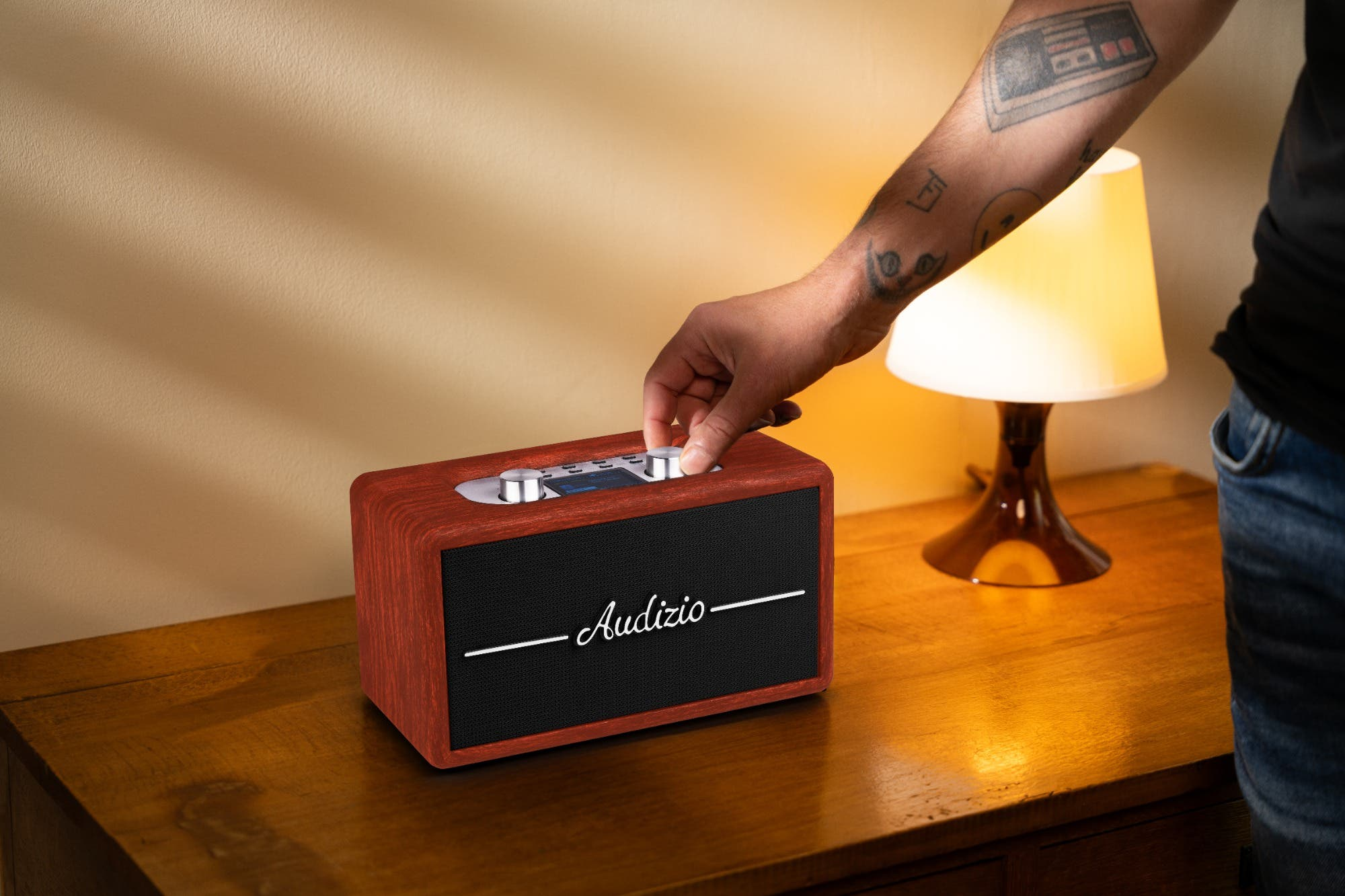 Una mano ajusta una radio de madera en una mesita de noche junto a una lámpara.
