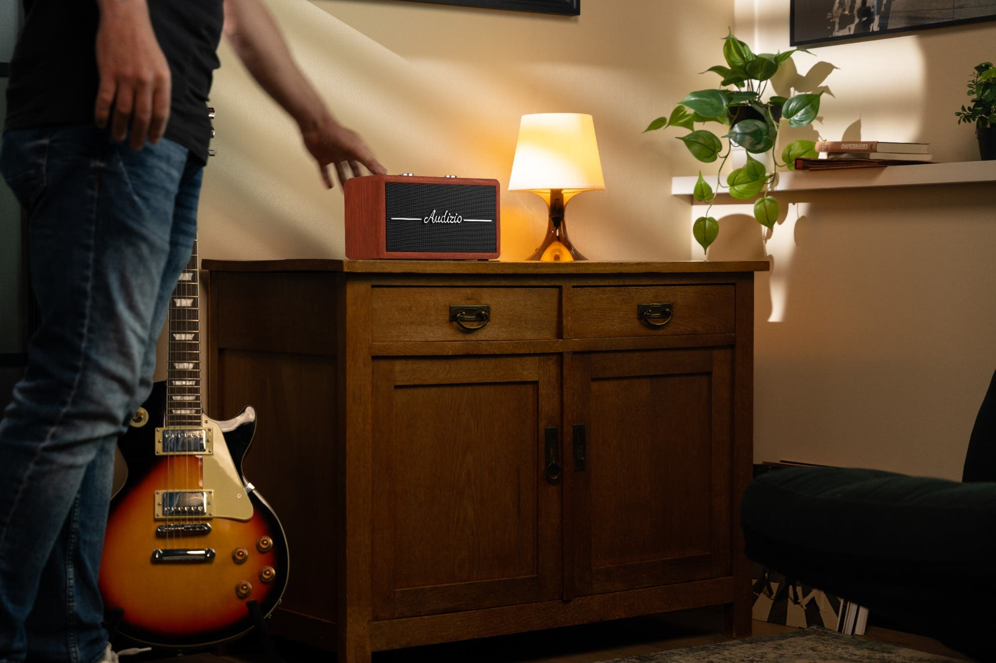 Un hombre toca un altavoz de madera junto a una lámpara en un mueble. Una guitarra eléctrica cerca.
