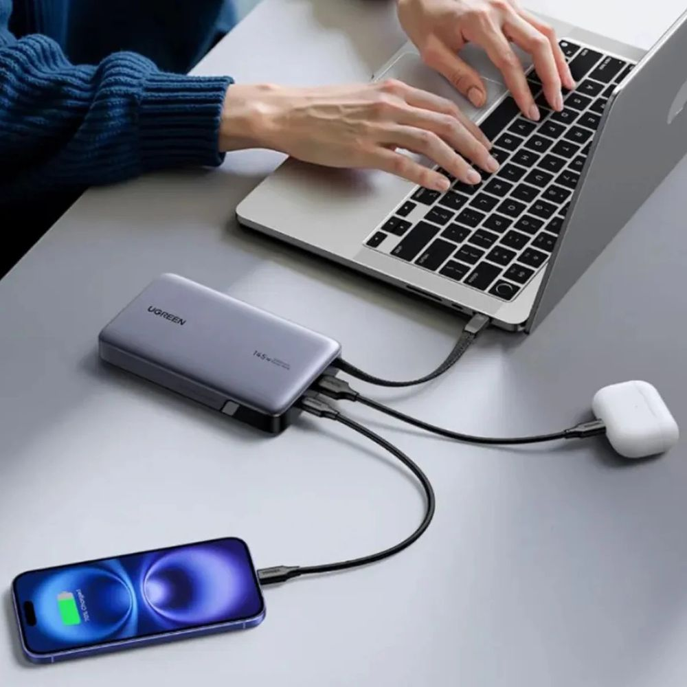 Szara powerbank ładuje laptopa, telefon i słuchawki, osoba pisze.